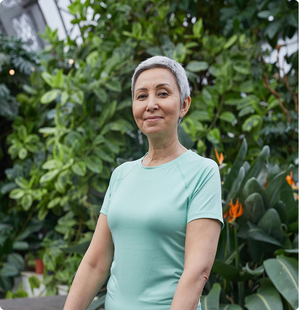 Portrait of Nalyn. She has short, gray hair, and she is wearing a blue shirt. A green bush is behind her.